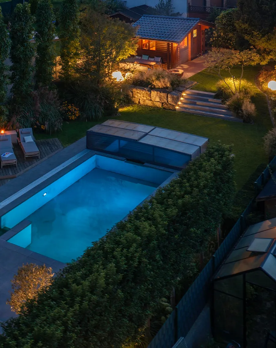 Luftaufnahme in der Dämmerung: beleuchteter Pool mit zurückgeschobener Überdachung, Rasen, Natursteinmauer mit beleuchteten Stufen, Leuchtkugeln, Säulenbäume und ein Gartenhaus mit Sitzplatz