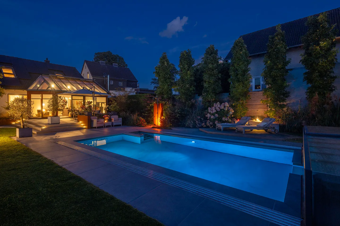 Von SURMANN gebauter Garten zur blauen Stunde: eine Reihe schmaler Bäume als Sichtschutz, Hortensien, Holzterrasse mit Liegen und beleuchteter Pool