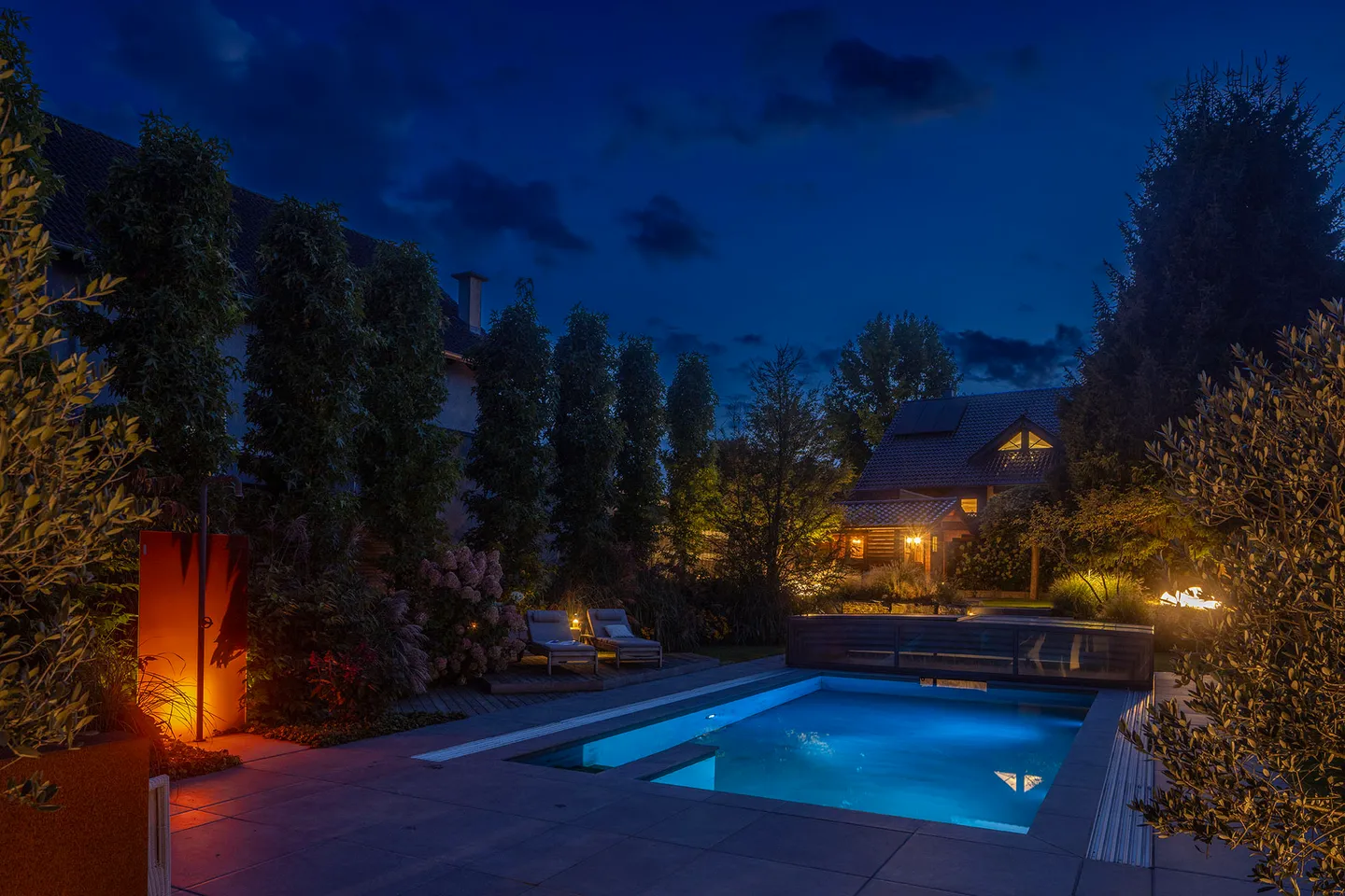 Garten bei Nacht: angestrahlte Cortenstahlwand mit Gartendusche, beleuchtete Bäume und Gräser, Liegen vor Hortensien und ein beleuchteter Pool