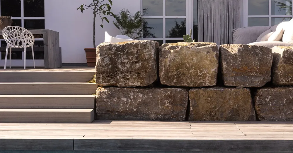 Natursteinmauer mit Treppenstufen und Holzdeck vor einem weißen Haus