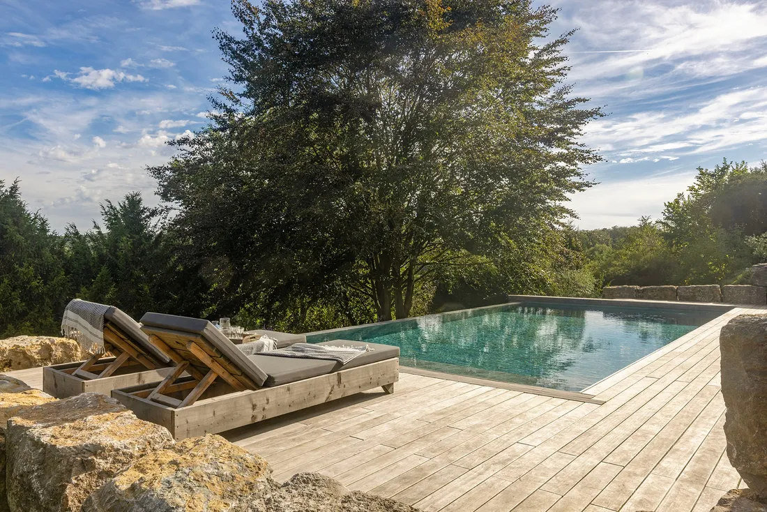 Pool mit Holzdeck und Natursteinmauer, dahinter Bäume und Blick in die Landschaft